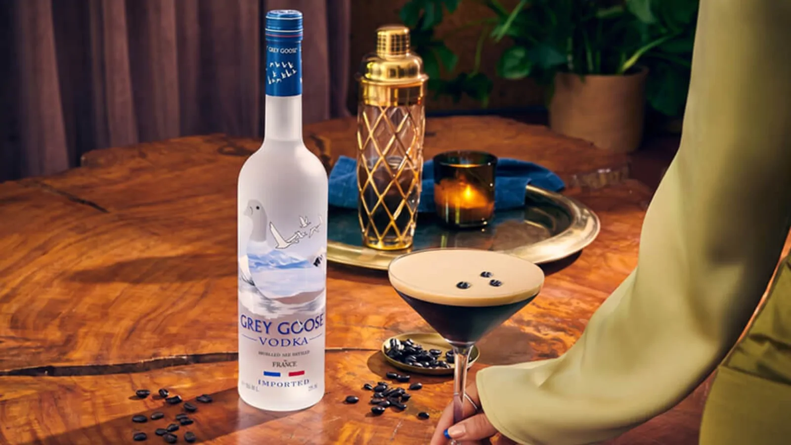 A bottle of Grey Goose vodka on a polished wooden table beside an espresso martini and cocktail shaker